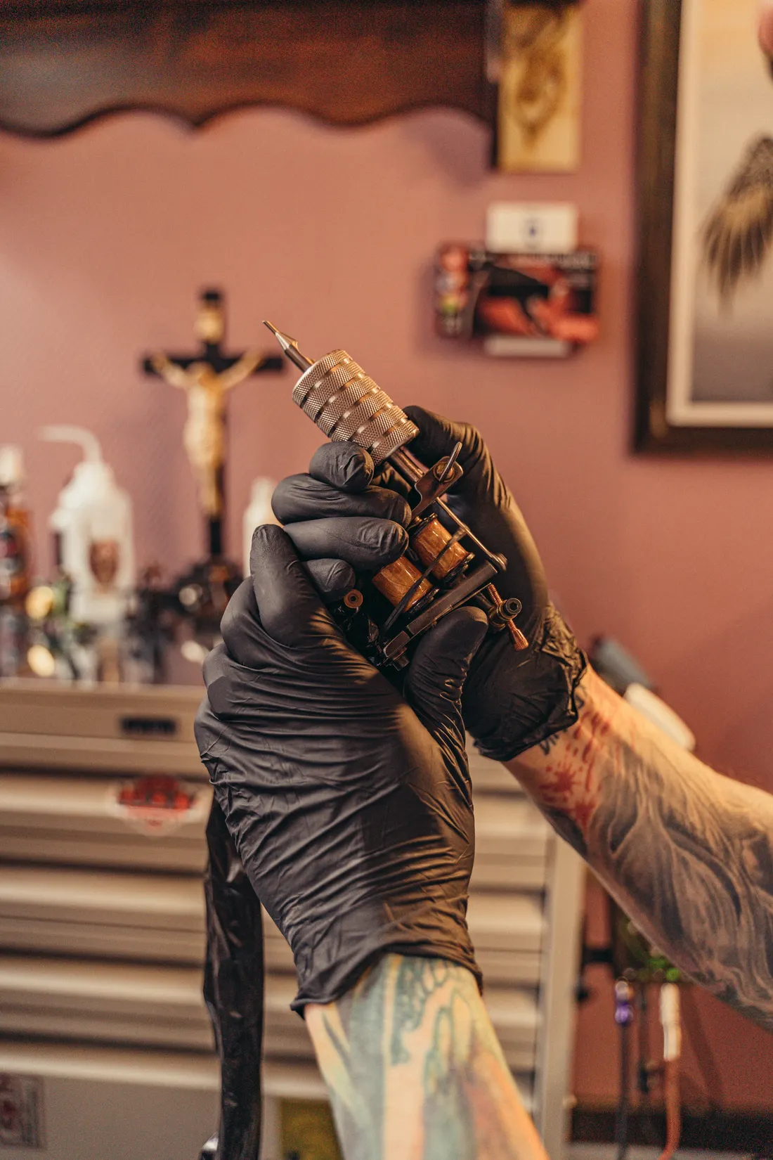 A tattooist's gloved hand holding a traditional coil tattoo machine in the studio